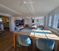 Bright living and dining area with wooden flooring, large windows, a sofa, a dining table with chairs, and a television on a stand.