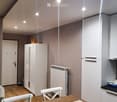 A modern, well-lit room with a wooden dining table and white chairs, a refrigerator, a cabinet, and a radiator. The room features a wooden floor and recessed ceiling lights.