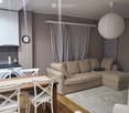 A modern, well-lit living room with a beige sectional sofa, a wooden dining table with white chairs, a white armchair, a washing machine, and a large round pendant light.
