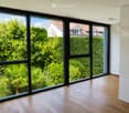 Empty room with wooden flooring and large black-framed windows offering a view of greenery and neighboring houses.