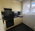 Modern kitchen with light-colored cabinets, black countertops, and stainless steel appliances including an oven, sink, and dishwasher. A window with blinds offers natural light.
