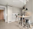 Modern kitchen with light grey cabinets, a wooden island, two black stools, and a sleek faucet. The space features pendant lights and a small decorative vase on the island.