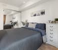 Modern bedroom with a large bed, nightstand, mirror, and decorative plants. The room features white walls, wooden flooring, and artwork on the walls.