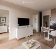 Modern, open-concept living and dining area with light wood flooring, white walls, and contemporary furniture including a sofa, coffee table, dining table with chairs, and a kitchen area with a sink and cabinets.