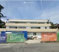 A two-story building with a flat roof and multiple windows, surrounded by construction banners and a partially visible construction site.