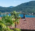 A scenic view from a rooftop terrace overlooking a lake surrounded by lush green hills and a small town. The foreground shows a tiled roof with palm plants, and the background features a calm lake with distant mountains.