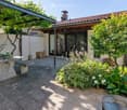 A well-maintained outdoor patio area with a stone table, benches, lush greenery, and a covered section leading to a house entrance.