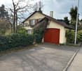2 story house, beige walls, red garage door, chimney, surrounded by trees and bushes