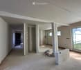 Empty room with unfinished concrete walls and floor, visible construction materials and tools, large window on one side, and open doorway leading to another room.