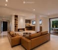 Modern living room with a large leather sofa, coffee table, fireplace, and open kitchen area with bar stools and a kitchen island. Shelves with decorative items are visible, and there is a dining area with a table and chairs through a sliding glass door.