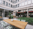 Courtyard seating area with wooden tables, green chairs, umbrellas, and potted plants, adjacent to a modern building with large windows.
