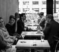 A group of people seated at tables in a restaurant or cafe, enjoying drinks and conversation. Tables are set with wine glasses, cups, and napkins.