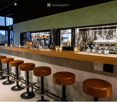 Modern bar area with a wooden counter, multiple bar stools, a refrigerator, a coffee machine, and a bowl of fruit on the counter.