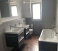 Modern bathroom with dark grey tiles, brown tiled floor, double sink vanity, bathtub, and large windows providing natural light.