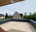 Modern rooftop terrace with beige tiled flooring, dark grey low walls, and a view of surrounding houses and greenery.