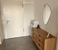 A small, neatly furnished room with a wooden dresser, a round mirror, and a small cabinet. The room has a tiled floor and white walls.