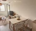 Bright, modern living and dining area with white walls, tiled floor, and large window. Features a sofa, coffee table, dining table with chairs, and a television on a stand.