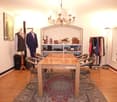 A well-lit room with a wooden table and chairs, a chandelier, shelves with various items, and mannequins dressed in suits.