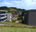 View of a modern residential complex with multiple buildings, including a multi-story apartment building with balconies, a smaller house with a red roof, and a contemporary structure with a dark facade. The area is surrounded by green hills and scattered