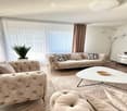 Modern living room with large windows, beige sofas, coffee table, ottoman, floor lamp, TV stand, and potted plant.