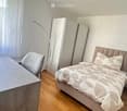Modern bedroom with a large bed, a wardrobe, a chair, a table, and a floor lamp. The room features wooden flooring and large windows with sheer curtains.