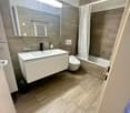 Modern bathroom with a white sink, large mirror, toilet, bathtub, and a white curtain. The floor is tiled in a light wood pattern, and the walls are covered in gray tiles.