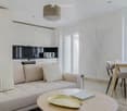 Modern, open-plan living and dining area with a light-colored sofa, coffee table, dining table with chairs, and a sleek kitchen area featuring a microwave and oven. The space is bright with large windows and a decorative wall design.