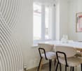 Modern dining area with a white dining table, four beige chairs, and decorative vases. Large windows provide natural light, and a framed abstract artwork adorns the wall.