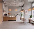 Modern, minimalist interior with pendant lights, white tables, chairs, a bench, and a counter with a coffee machine and display case.