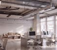 Modern office space with exposed ceiling beams, large windows, desks, chairs, a sofa, coffee table, and computer monitors.