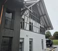 Modern two-story house with white and black exterior, large windows, and a distinctive roof design. The ground floor features a covered entrance area with a paved walkway.