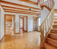 Spacious hallway with wooden flooring, exposed wooden beams on the ceiling, and a wooden staircase with a carved railing leading upwards. Several doors open to adjacent rooms, including a kitchen area visible in the background.