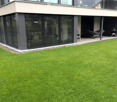Modern house exterior with a well-maintained lawn, a covered patio area, and outdoor furniture including a hammock and grill.