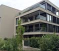Modern multi-story residential building with large glass balconies and contemporary design, surrounded by lush greenery.
