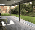 Covered patio with tiled floor, glass walls, outdoor furniture including a table, chairs, and a grill, overlooking a green lawn and neighboring houses.