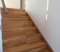 Wooden staircase with a handrail, leading up to an open doorway, with white walls and a slanted ceiling.