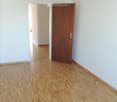 Empty room with light-colored wooden flooring and white walls. A wooden door is visible on the right side, and an open doorway leads to another room in the background.