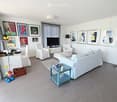 Bright living room with white walls, grey tiled floor, large sofa, coffee table, armchairs, ottoman, TV, and decorative artwork.
