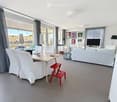 Open-plan living and dining area with large windows offering a view, featuring a white sofa, dining table with chairs, a red stool, and a television.