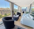 A modern, furnished balcony with a white sofa, two black armchairs, a small table, and a grey ottoman. The space features large glass windows and a view of the city.