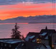 Panoramic view of a lake and snow-capped mountains at sunset, with rooftops of houses in the foreground.