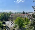A panoramic view of a residential area with multiple buildings, trees, and a clear blue sky. The scene includes a mix of modern and traditional houses, surrounded by greenery and pathways.