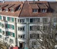 Multi-story residential building with red-tiled roof, white walls, green shutters, and multiple windows. The structure features a mix of architectural styles with a prominent curved section and several dormer windows on the roof.