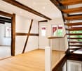 Empty room with wooden floor, exposed wooden beams, and a staircase leading upwards. The space is well-lit with recessed lighting and has white walls with a textured finish. There are windows on one side and a door leading to another area.