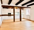 Empty room with wooden floor, white walls, exposed wooden beams, white kitchen cabinets, and a window with a view of a tiled roof.