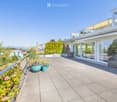 Balcony with modern glass railings, outdoor seating, and potted plants, overlooking a scenic landscape with trees and distant buildings.