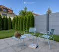 A well-maintained garden area with a light blue metal table and two matching chairs, a small round table with a vase and a cup, and a stack of books on the ground. The garden is enclosed by a grey wooden fence and features neatly trimmed green hedges and