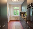 Modern kitchen with dark grey cabinets, stainless steel appliances, a central island, and a large window overlooking greenery. Features a round ceiling light fixture, a radiator below the window, and a textured rug on the terracotta floor.