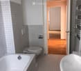 Modern bathroom with a bathtub, toilet, sink, and radiator. The walls are tiled, and there is an open doorway leading to another room with wooden flooring.
