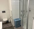 Modern bathroom with white tiled walls, a glass-enclosed shower, toilet, toilet paper holder, and a blue laundry basket on a rug.